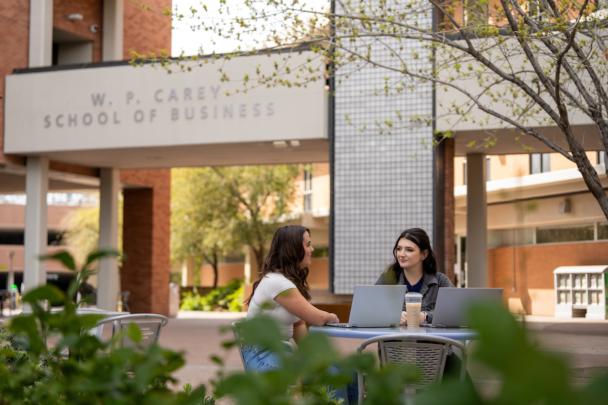 Two ASU students sitting at an outside table near the W. P. Carey School of Business buildings