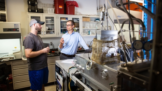 Arizona State University Assistant Professor, Zachary Holman (right), talks with Doctoral Student, Peter Firth, next to a prototype of a nano-particle deposition tool. Holman was awarded a Moore Inventor Fellowship to further develop a technique that can add beneficial properties to ordinary surfaces through nano-particle coatings. Photographer: Deanna Dent/ASU