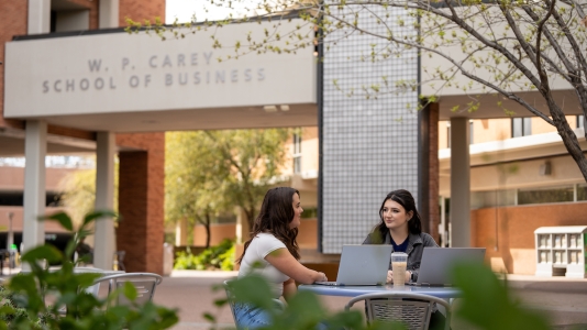 Two ASU students sitting at an outside table near the W. P. Carey School of Business buildings